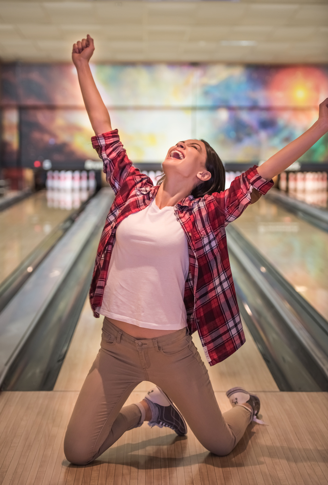 Girl at the bowling alley, in front of a lane on her knees. Her arms are extended in the air showing excitement, showing personality.