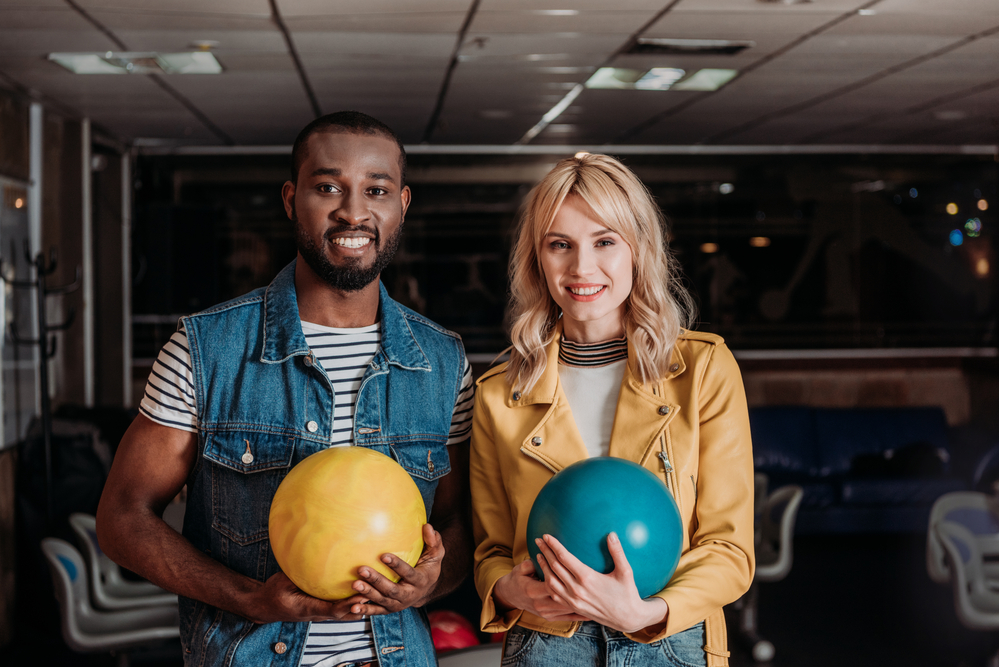 Young man and woman standing in a bowling alley. They are both holding bowling balls in their hands.