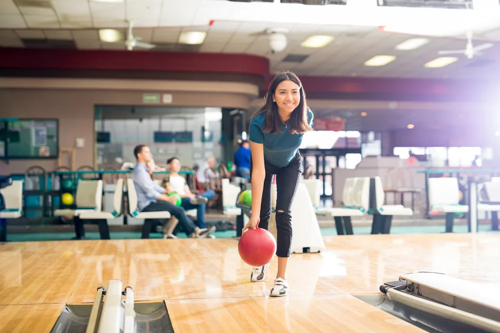 Full length of smiling teenage girl throwing ball while training for bowling in club. A good bowling score for age 13 to 19 is 120 to 190 points.