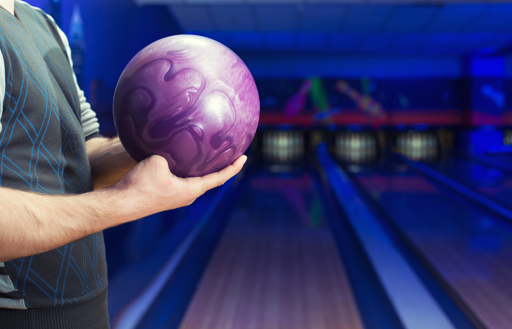 A man holding a purple bowling ball with a conventional grip standing inside of a local bowling alley.