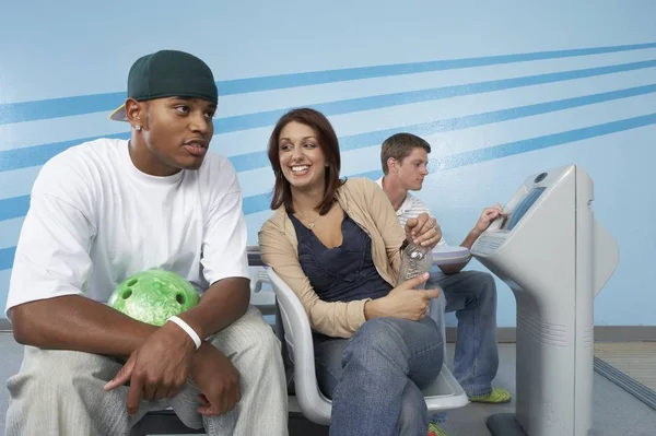 This image is of 3 bowlers that are distracted. One guy is chatting while holding his bowling ball. The guy is sitting chatting and the third person is looking at the scoring console.