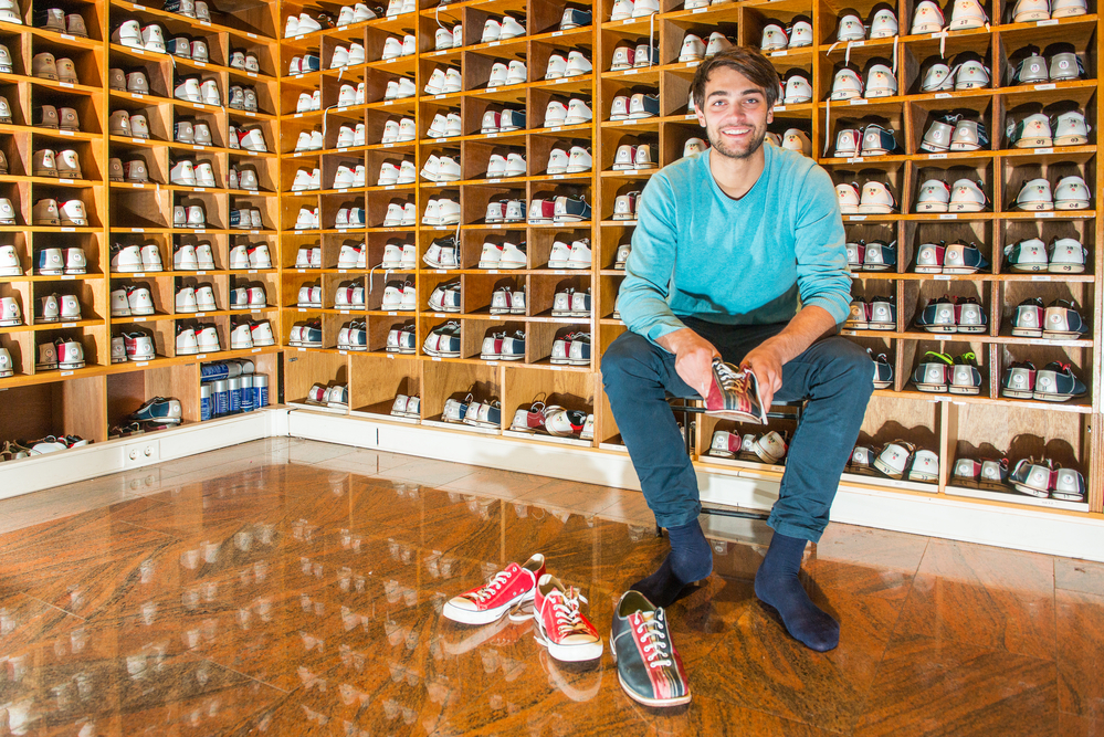 A man is shoe shopping (or choosing shoes) as he tries to find the right pair of sliding shoes for wooden bowling alleys.