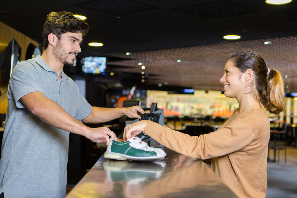 A lady bowler renting shoes from the counter is surprised that bowling is such an expensive sport for casual players.
