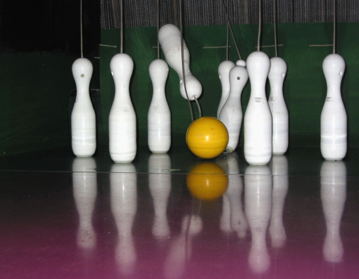 This image is of a game of nine-pin bowling. A yellow ball is rolled towards the pins.
