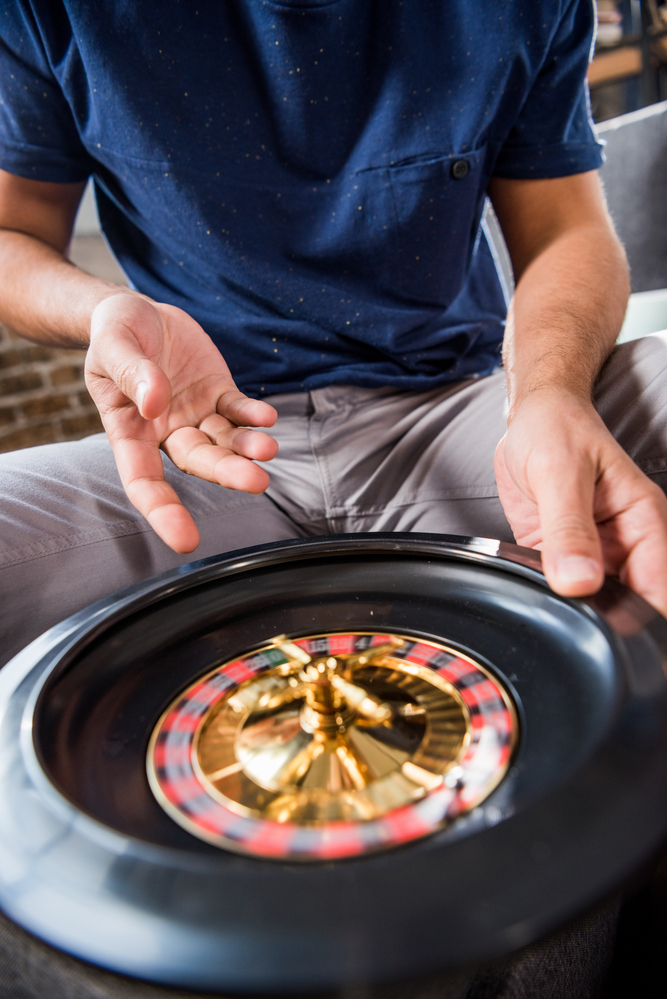 A guy in a blue shirt sitting at a table with the small roulette wheel. It's his turn to spin after bowling his two frames.