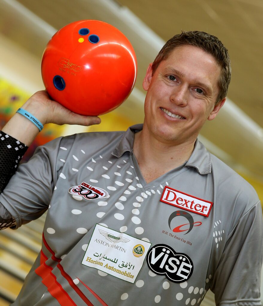 Professional bowler in a bowling shirt with fun colors, holding a red bowling ball.