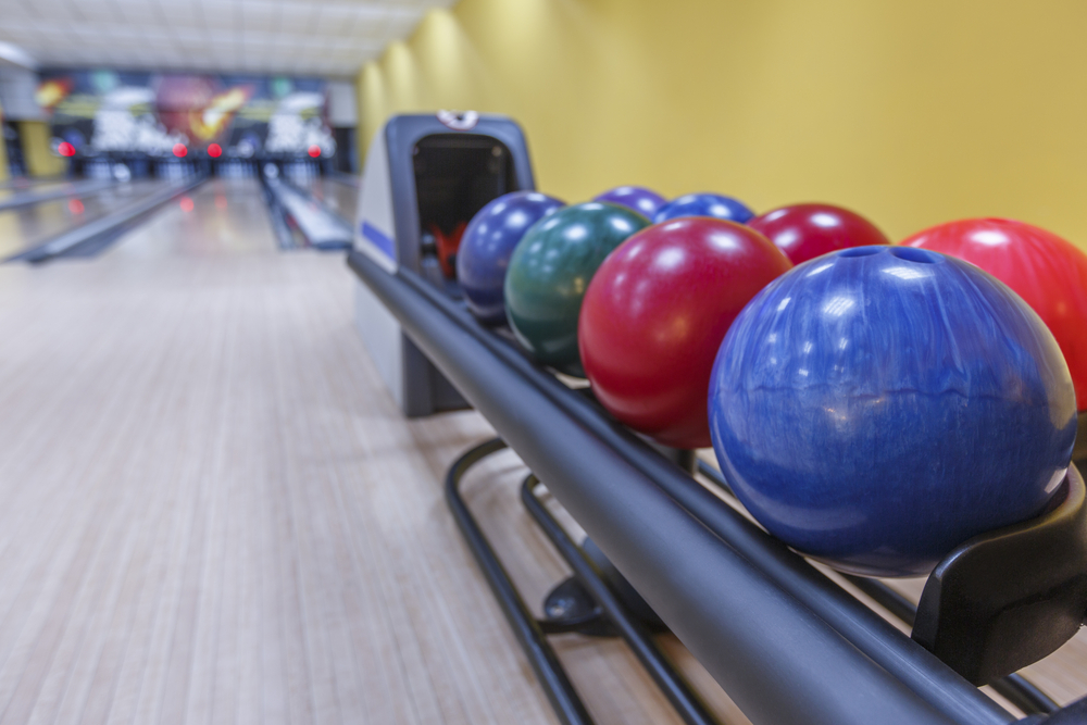 On a ball return rack, next to a yellow wall, are types of bowling balls varying from a custom bowling ball, urethane bowling ball or particle bowling balls.