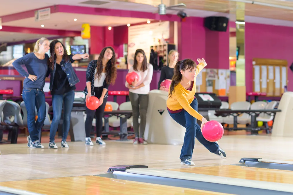 Six ladies are on a lane, three of them are holding bowling balls and wearing denim jeans, jackets, and all wear bowling shoes because it is mandatory.