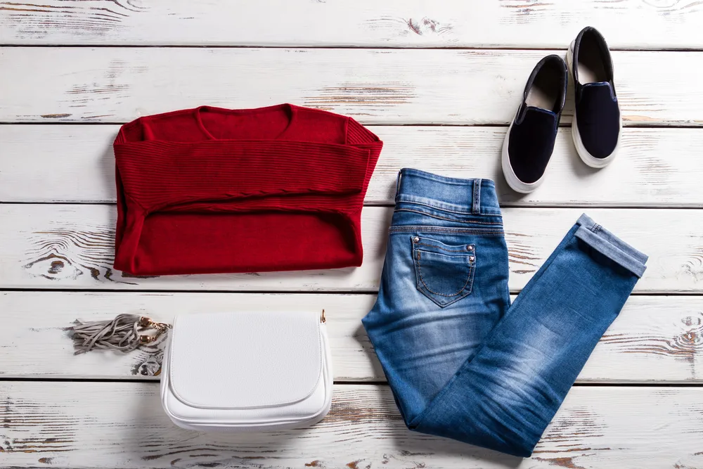 Red sweater, black shoes, high waisted jeans, rolled up jeans at the cuffs, and white clutch bag on a white wood background.