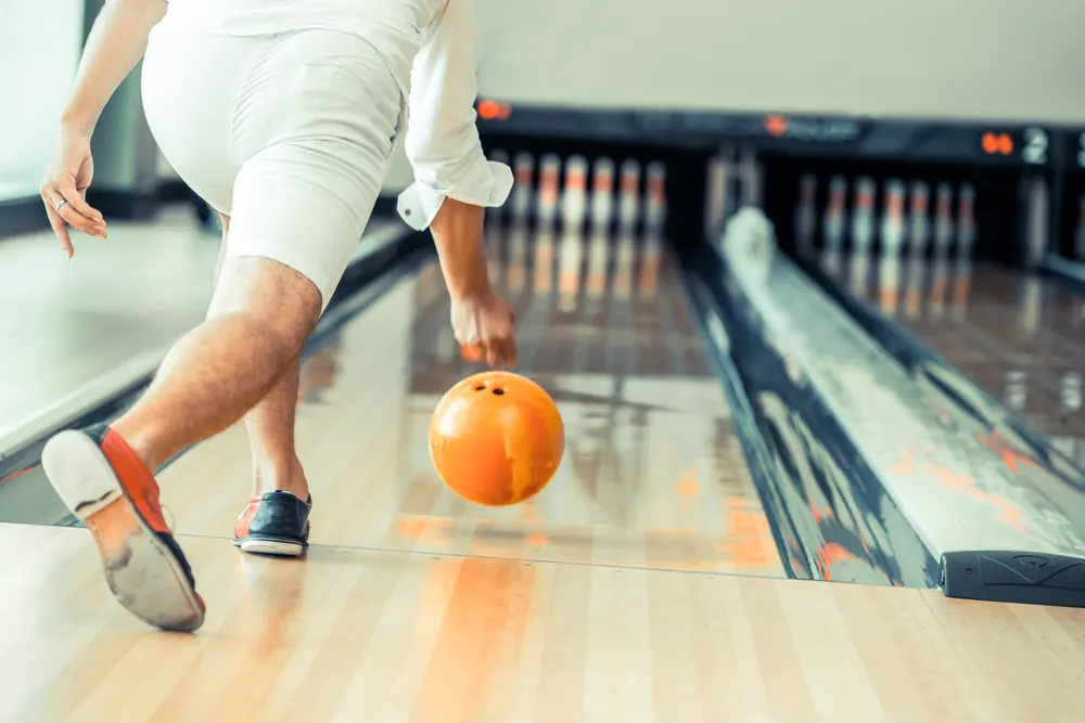 Right handed bowler wearing tri-colored bowling shoes, approaches the foul line, uses a bowling technique for a hook shot, using a urethane or resin coating orange bowling ball.