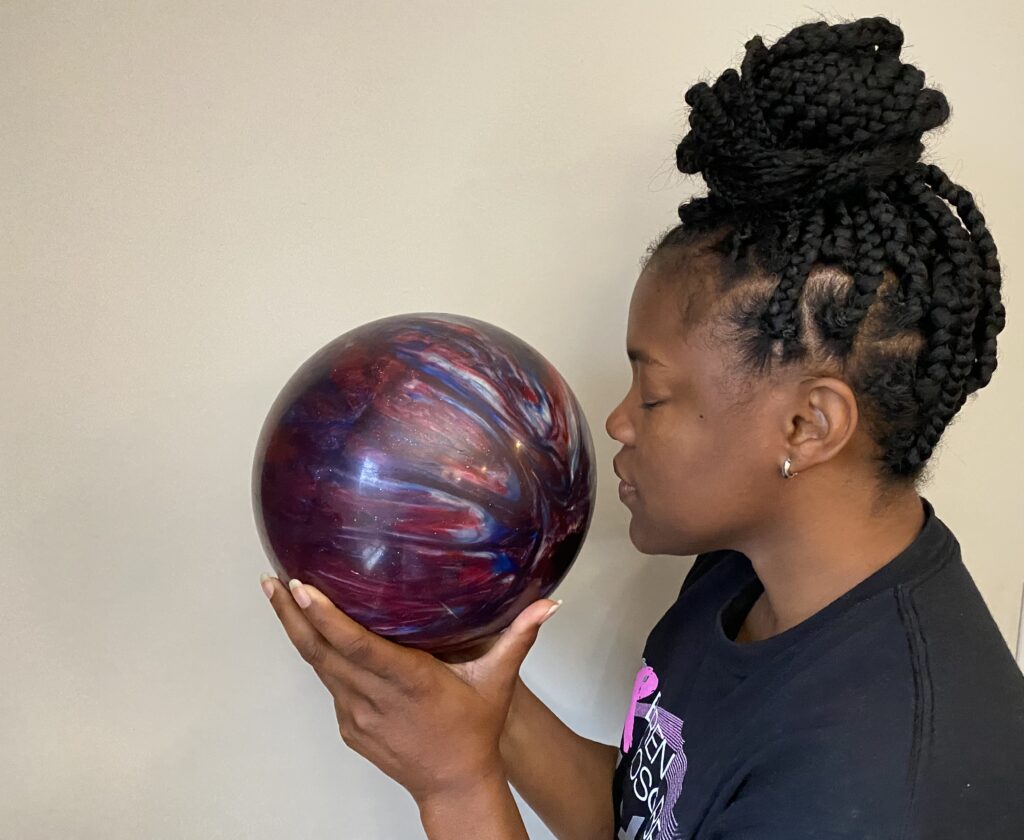 Knowing storm balls are scented, a woman in a black shirt sniffs a red, blue, and white swirled bowling ball.
