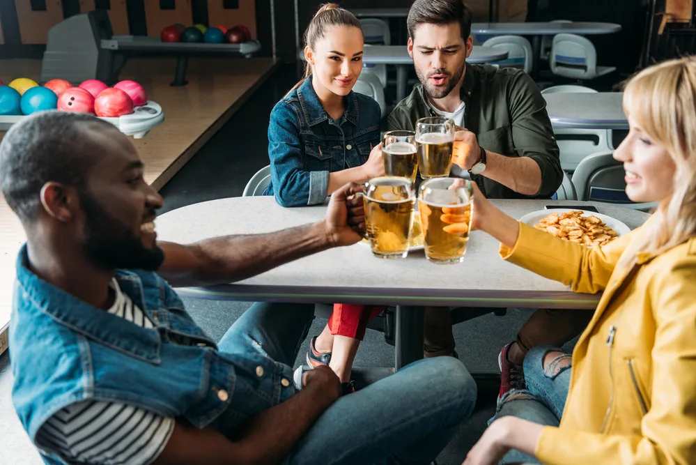 Friends sitting around a bowling table at the bowling club, each of them has a miniature pitcher of beer in their hand. They wanted to play beer bongs but opted instead for beer bowling.