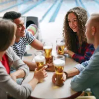 Friends sitting a table at a bowling alley, near a bowling ball return; thinking how about a bowling drinking game that involves making two strikes.