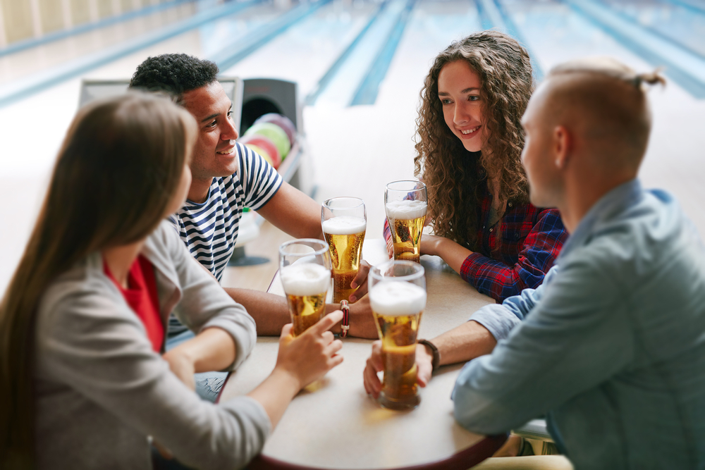 Friends sitting a table at a bowling alley, near a bowling ball return; thinking how about a bowling drinking game that involves making two strikes.