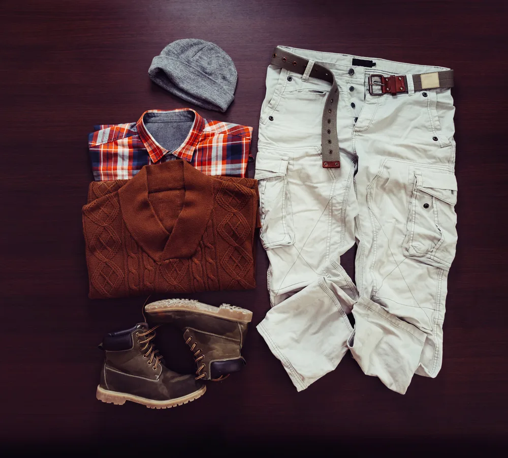 Gray skull cap, burnt red cardigan, plaid colored button-down shirt, brown boot,s and comfy tan cargo pants on a rich brown background.