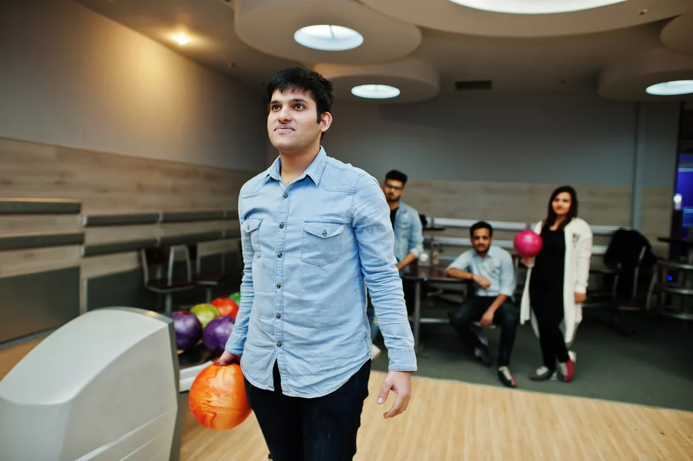 The man in the blue shirt stands at the line with an orange bowling ball to put into the physical game that the bowling instructor showed him.