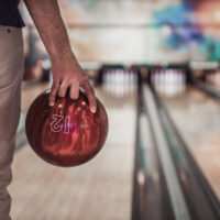 The male bowler is standing in the approach area set to use the straight bowling technique with red 12 pound bowling ball.