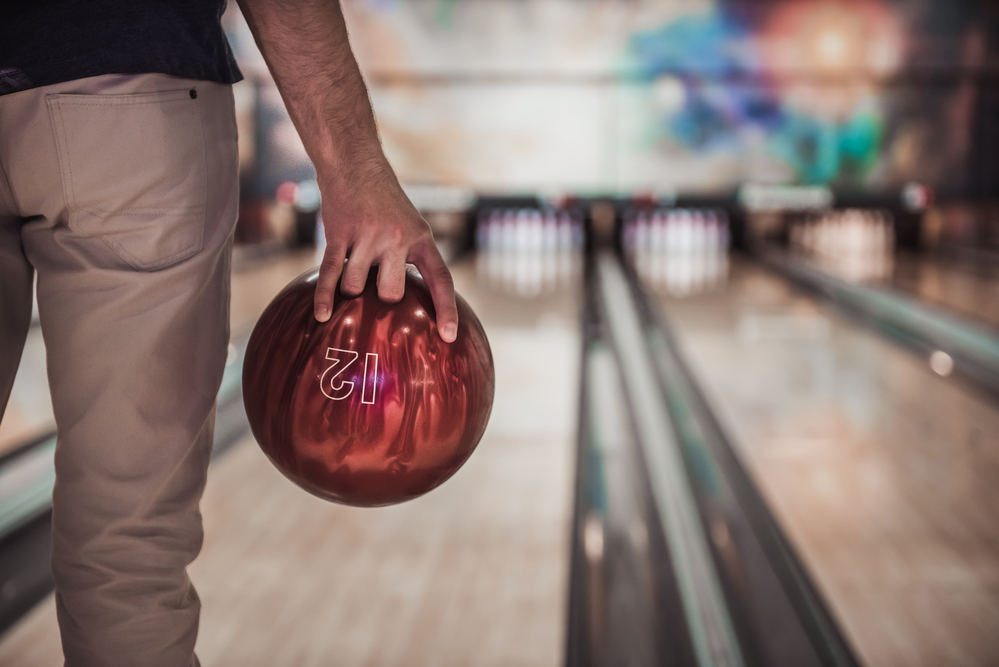 The male bowler is standing in the approach area set to use the straight bowling technique with red 12 pound bowling ball.