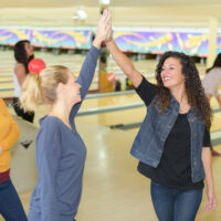 Three female friends high five each other at the bowling center after playing a game. The friend in the yellow shirt is holding a bowling ball.