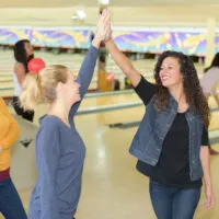 Three female friends high five each other at the bowling center after playing a game. The friend in the yellow shirt is holding a bowling ball.