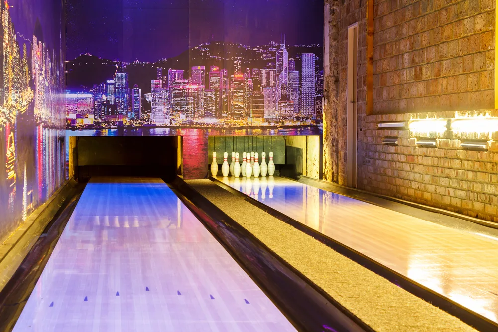 Two lane bowling alley with pins set on one side and pins were knocked down on the other lane with a backdrop of the city on the walls.
