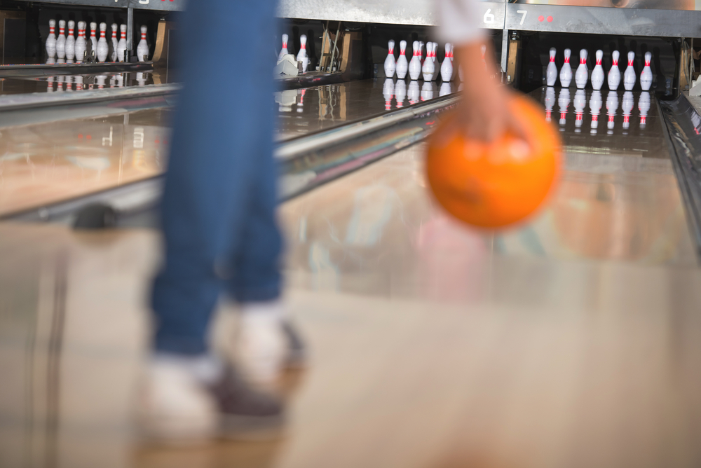 The female bowler standing in the approach area with an orange bowling ball is set to use the backup bowling technique for the strike.