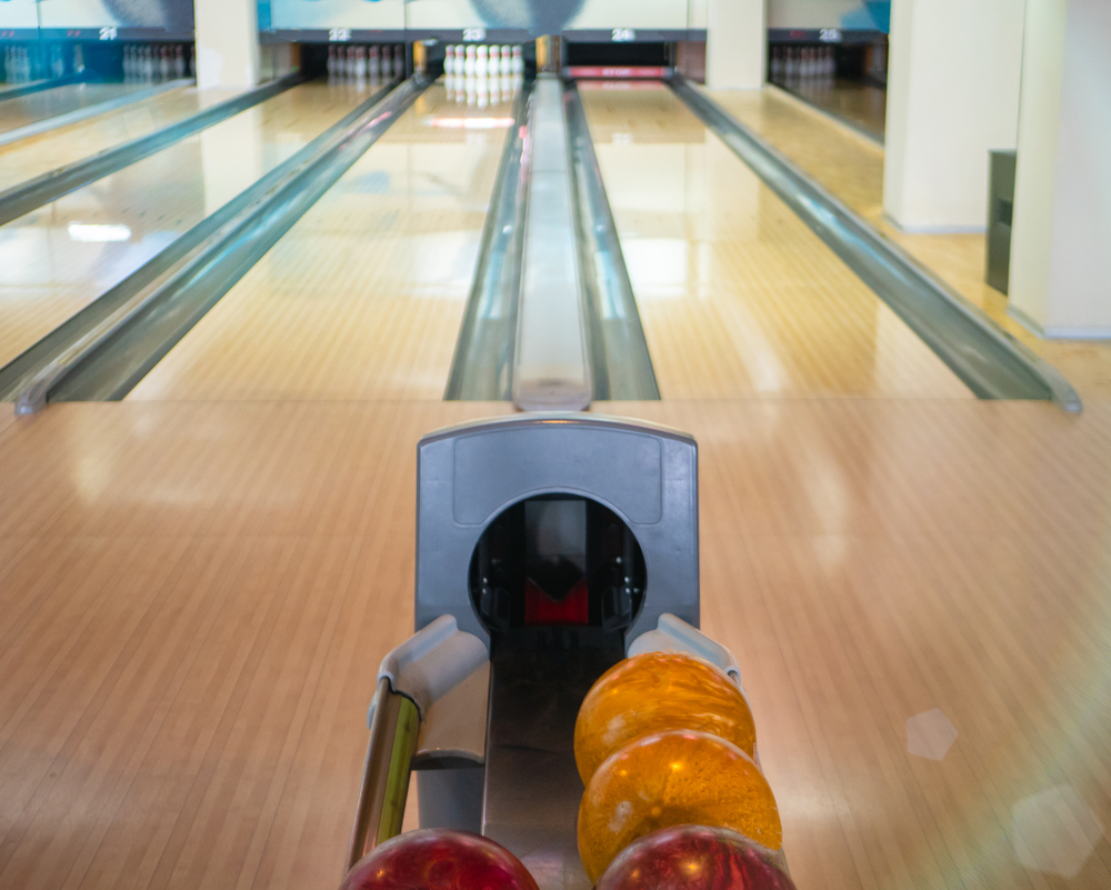 Seven lanes displayed with a focus point on a ball return, and four bowling balls are visible. Due to the sport oil pattern, the bowler has to decide which ball to use for a strike during sport bowling.