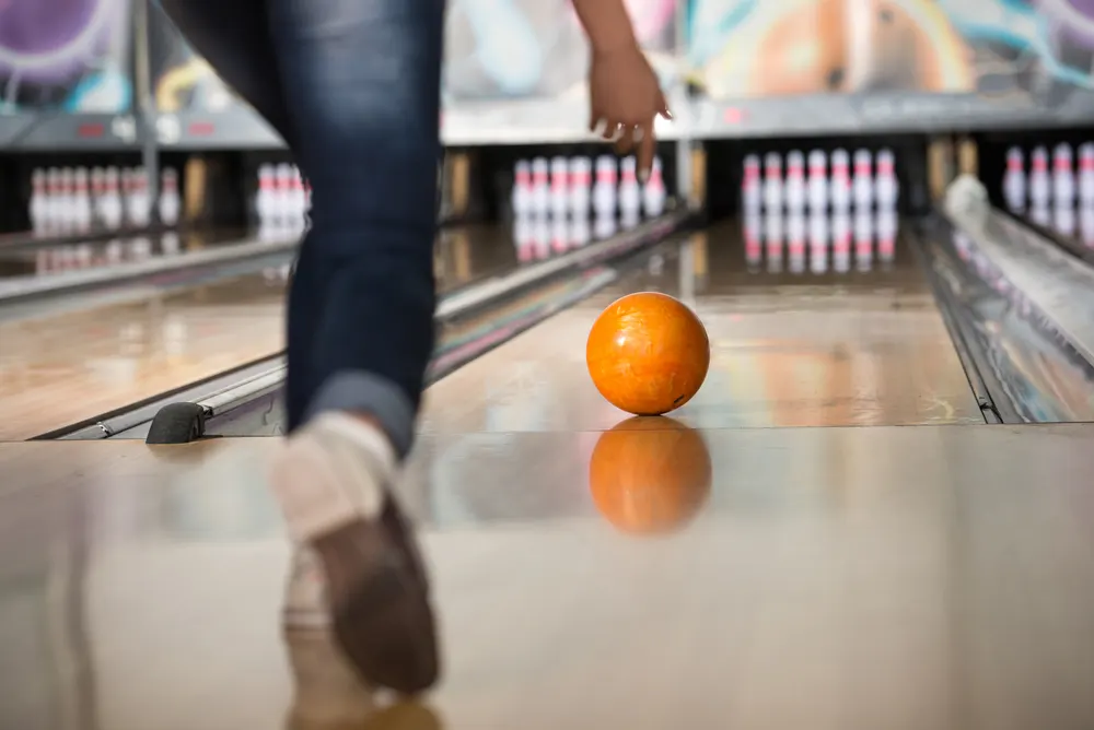 Young lady releases the orange bowling ball at the foul line and it rolls down the lane.