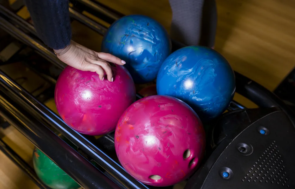The woman picks up the ball from the other bowling equipment and stands in front of the bowling lanes.