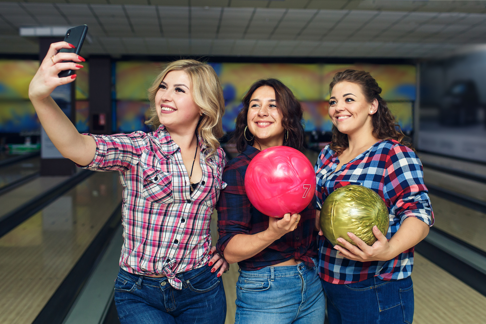 Girlfriends take selfies with smartphone at the bowling alley wearing crop tops and high waisted jeans.