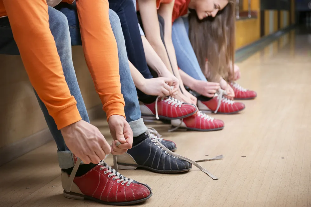 A girl with long hair, wearing a graphic tee is lacing up comfy rental bowling shoes.