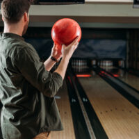 Bowler standing at foul line holding a red bowling ball with fingers and thumb inserted with a stiff wrist.