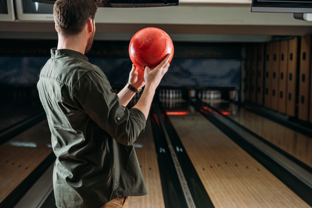 Bowler standing at foul line holding a red bowling ball with fingers and thumb inserted with a stiff wrist.
