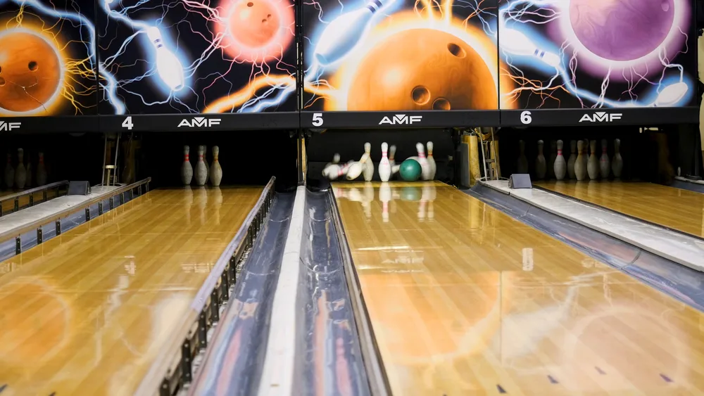 A bowling alley with wooden floors focused on two lanes where the throw will likely leave the 1 and 2 pins.