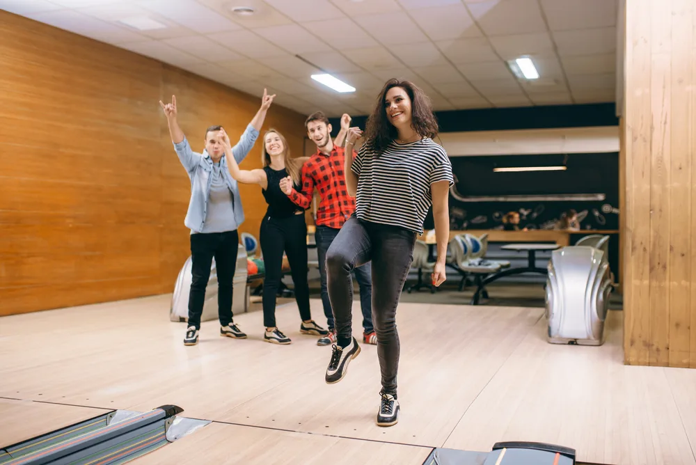 A female bowler standing near a ball return just threw a strike while his friends celebrate in the background.