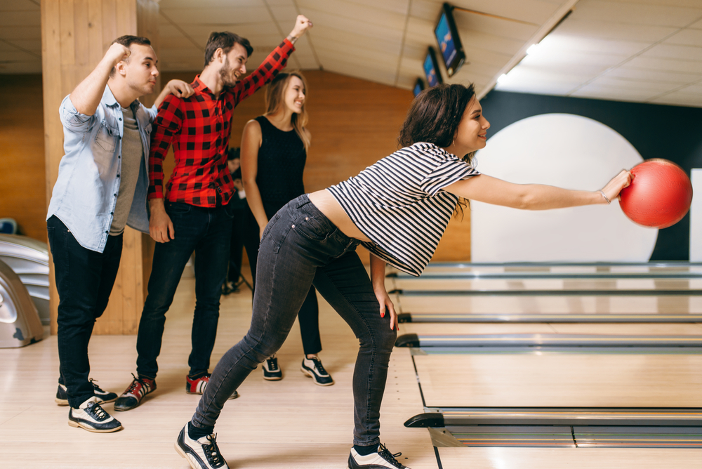 A female bowler with a family logo in the background throws the ball with incorrect technique while avoiding a gutter.
