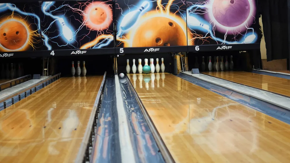 A light blue colorful bowling ball hits the middle pin on wooden pine floors with a playful graphic-filled background.