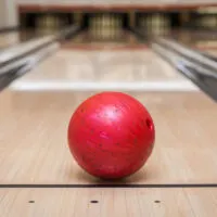 The red ball rolls down the lane with the finger holes and thumb holes visible following an open frame on a double wood lane.