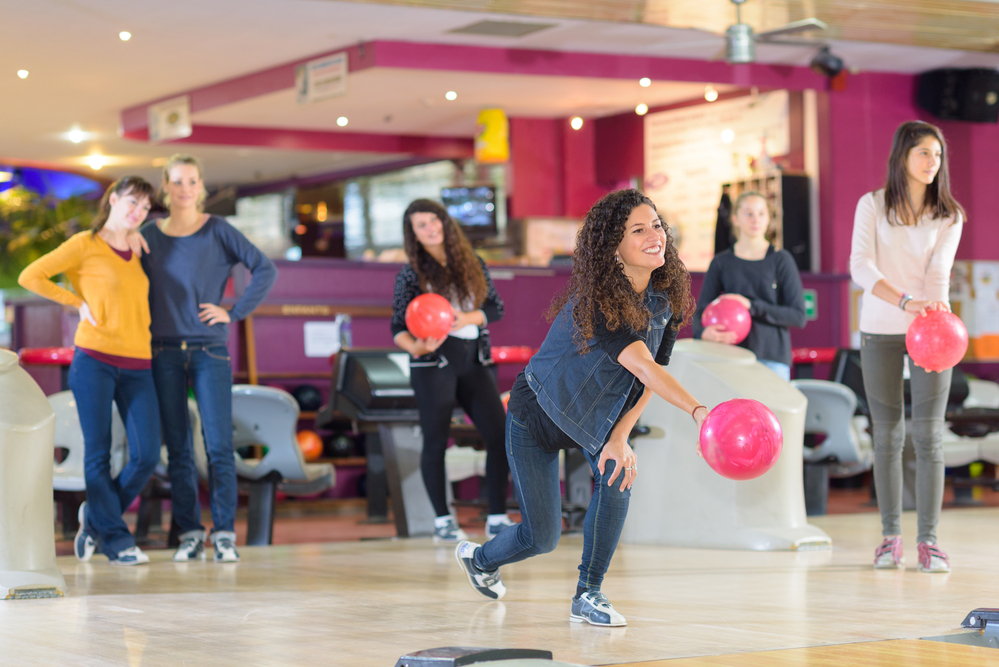 Six ladies bowling together and the lady in the denim jacket is rolling a pink bowling ball.