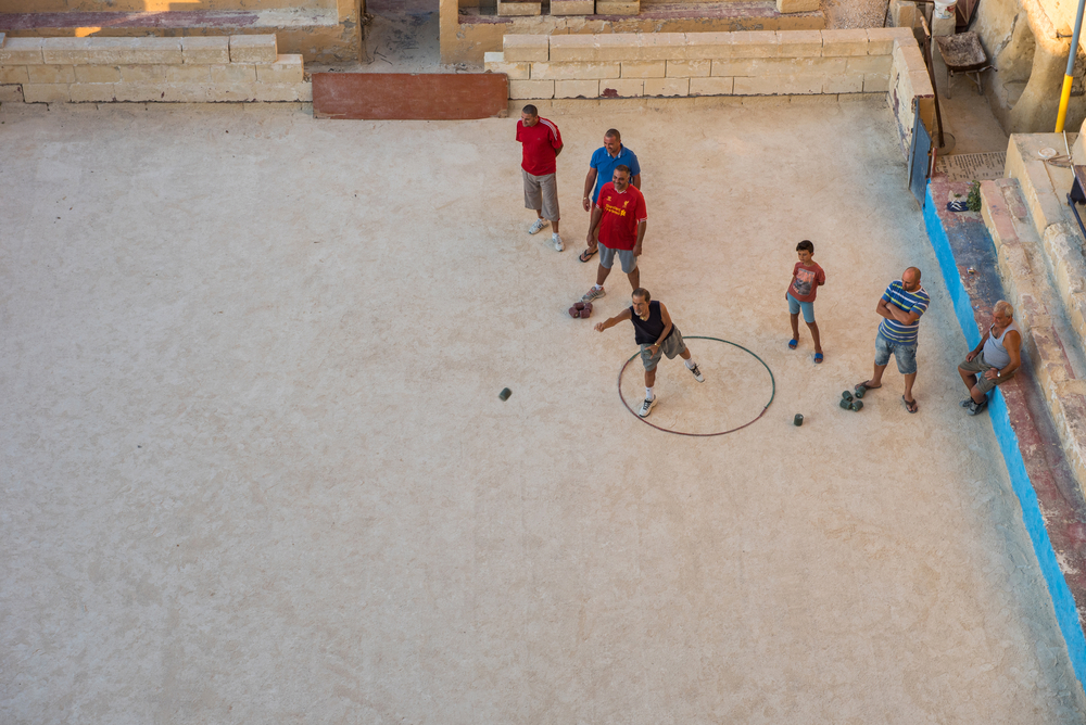 Seven men are playing on a hard-packed dirt surface, the most for bocce courts.