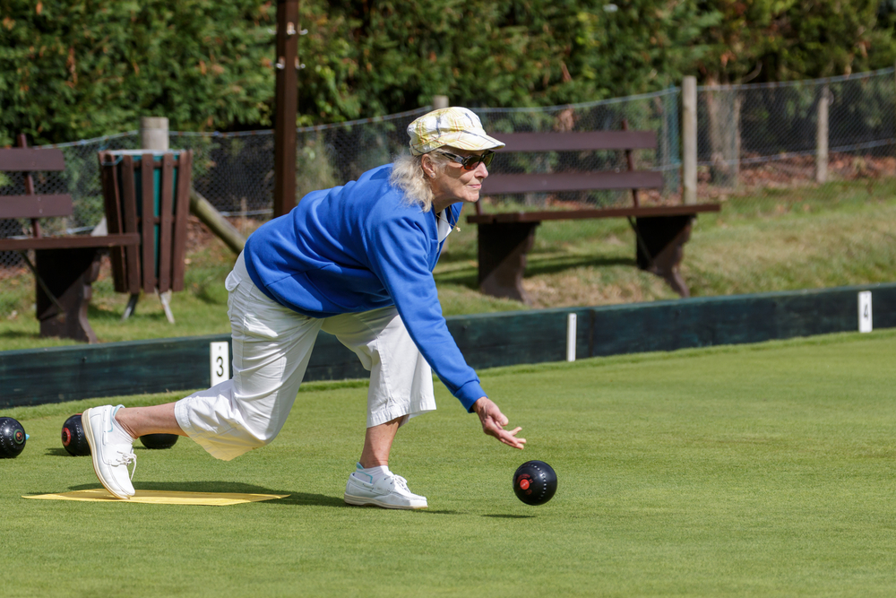 A lady in a blue sweater and hat is playing on the lawn bowling rink, which is bent grass and is usually 19 feet by 120 feet.