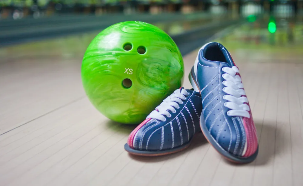 A green typical ball layout with two finger holes and a thumb hole, found at bowling centers, next to blue and red bowling shoes.