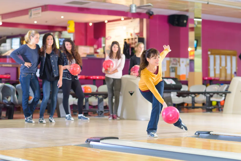 The woman bowler with the yellow shirt and pink ball, left knee is bent slightly as she releases the ball.