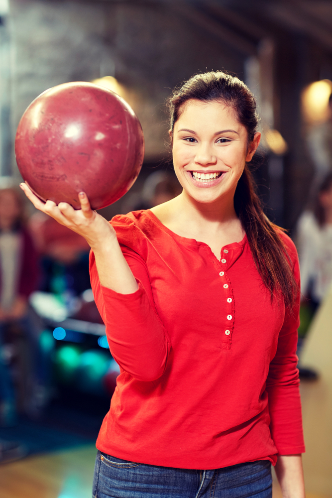 Female bowler in bowling alley holding red bowling ball smiling, had a perfect swing and got a strike