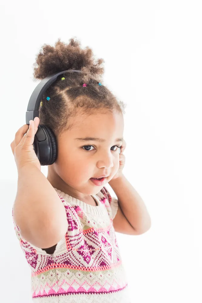 Little girl in a colorful dress listening to music with headphones on a white background because many parents are aware of loud noises at local bowling alley