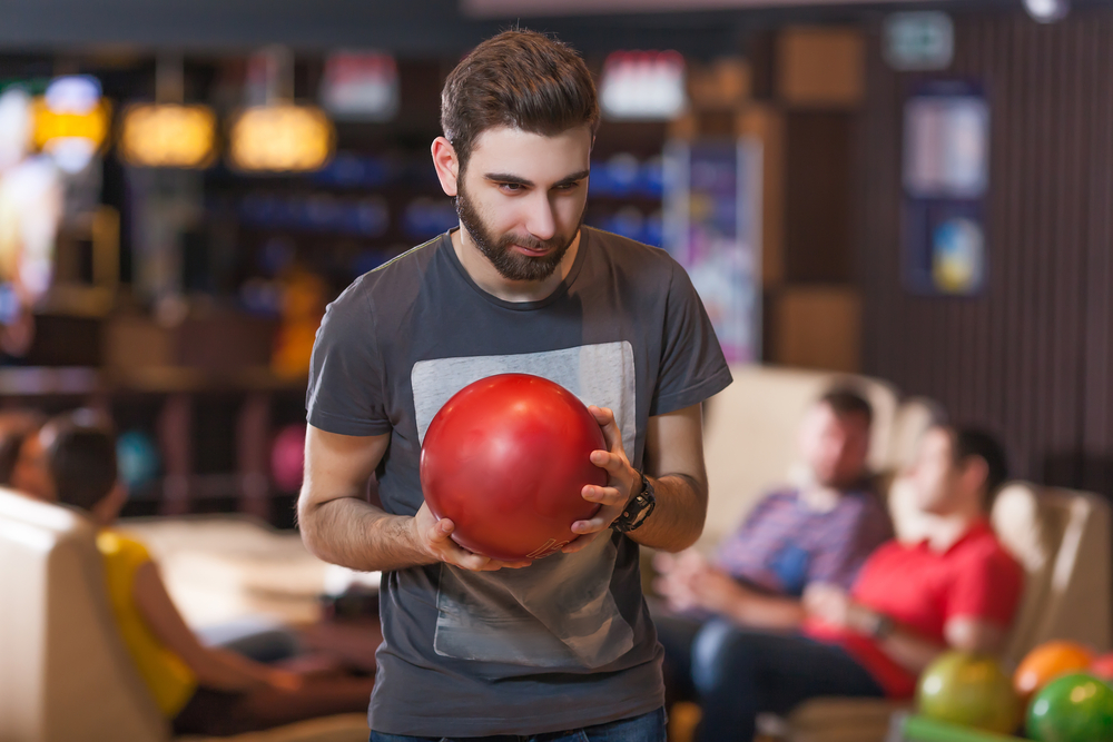 Bearded man with red bowling ball at bowling alley has relevant concern the difficult of the pattern will affect his average
