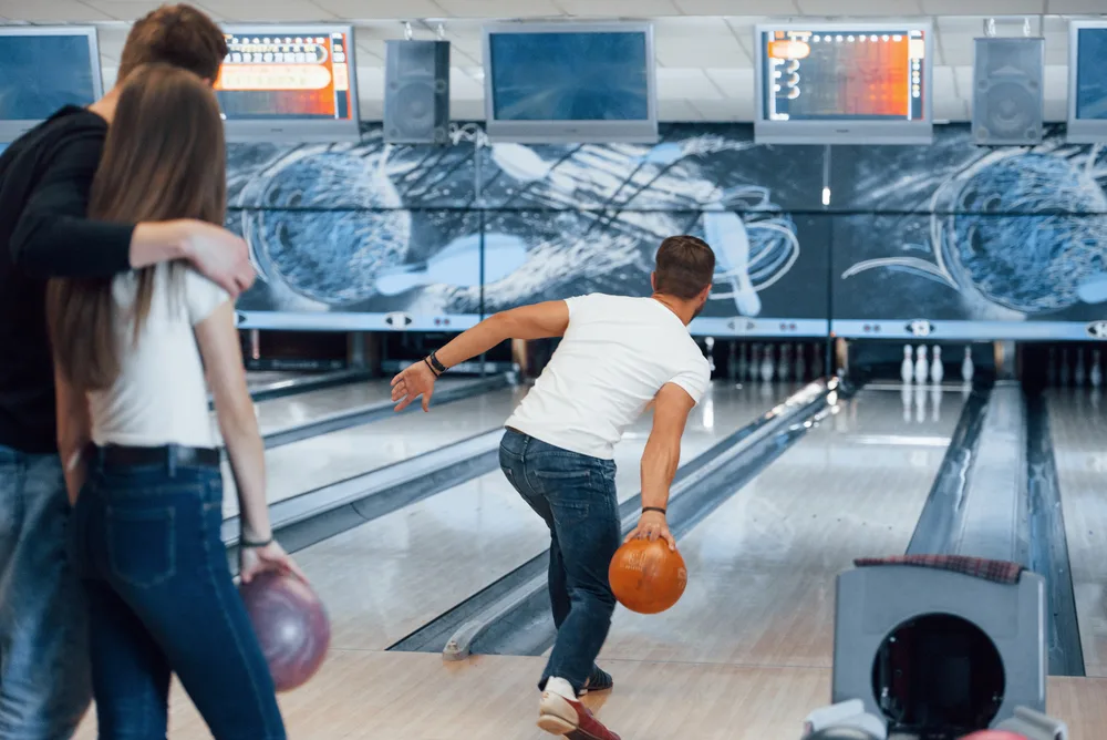 Bowler in white tee shirt roll the orange colored ball with his bowling arm.