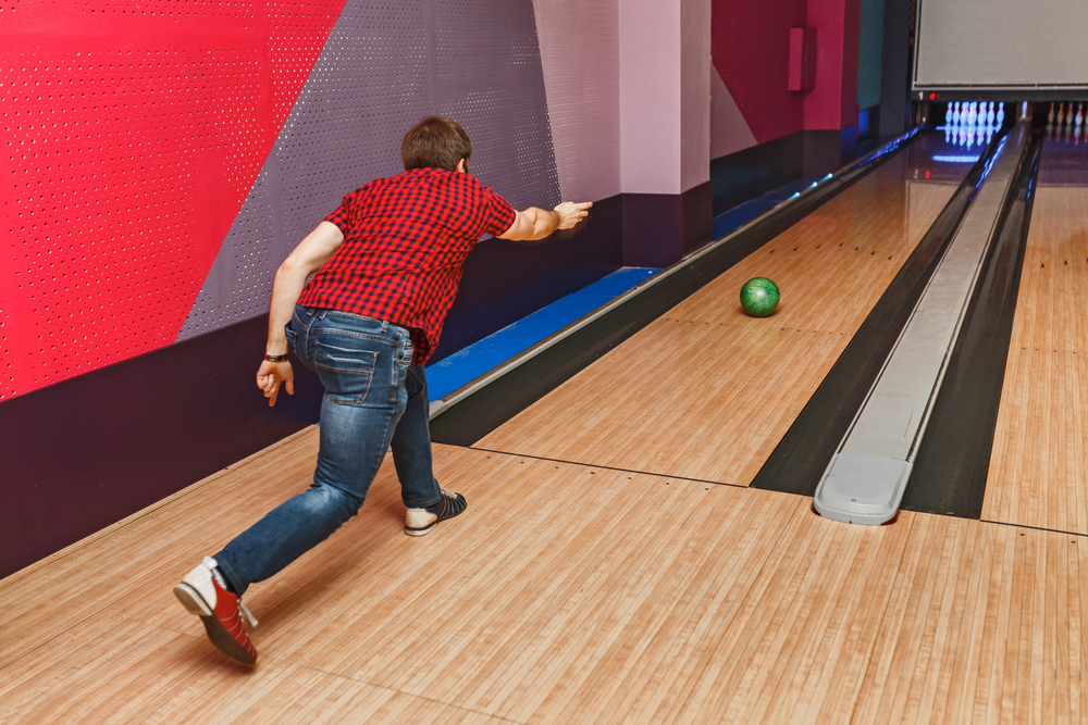 Young man in a checkered shirt throwing green bowling ball down alley to earn 10 points scored for a strike.