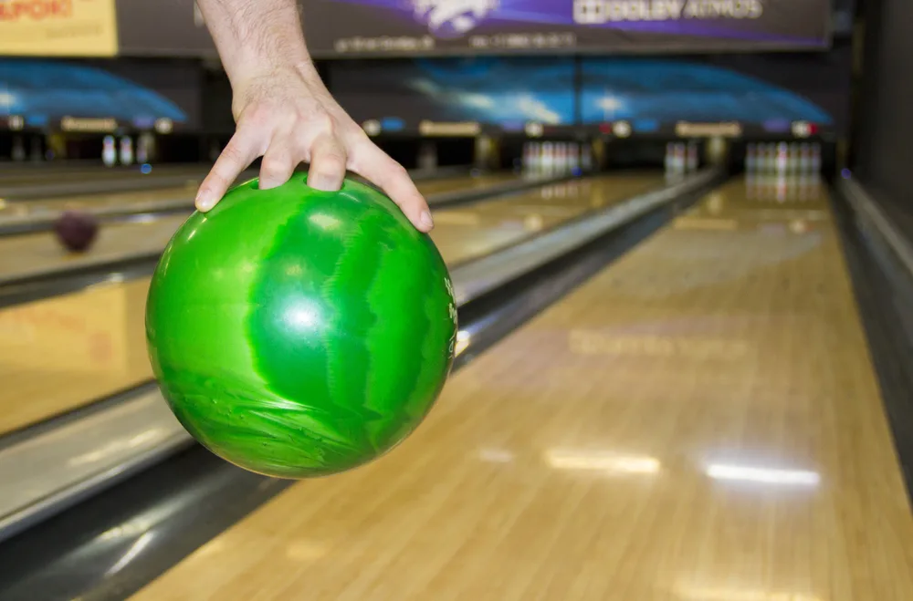 A man's hand throws a green bowling ball on a lane that has too much oil, the excess oil will affect his game