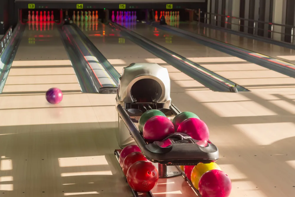 Playing area with 16 lanes with bowling pins, a bowling ball rolling along the lane, and colored bowling balls in the pit in the modern pin bowling alley